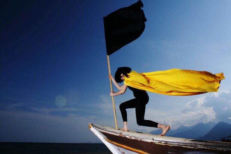 A woman in a bold pose with a black flag and yellow cape on a boat by the sea under blue skies.