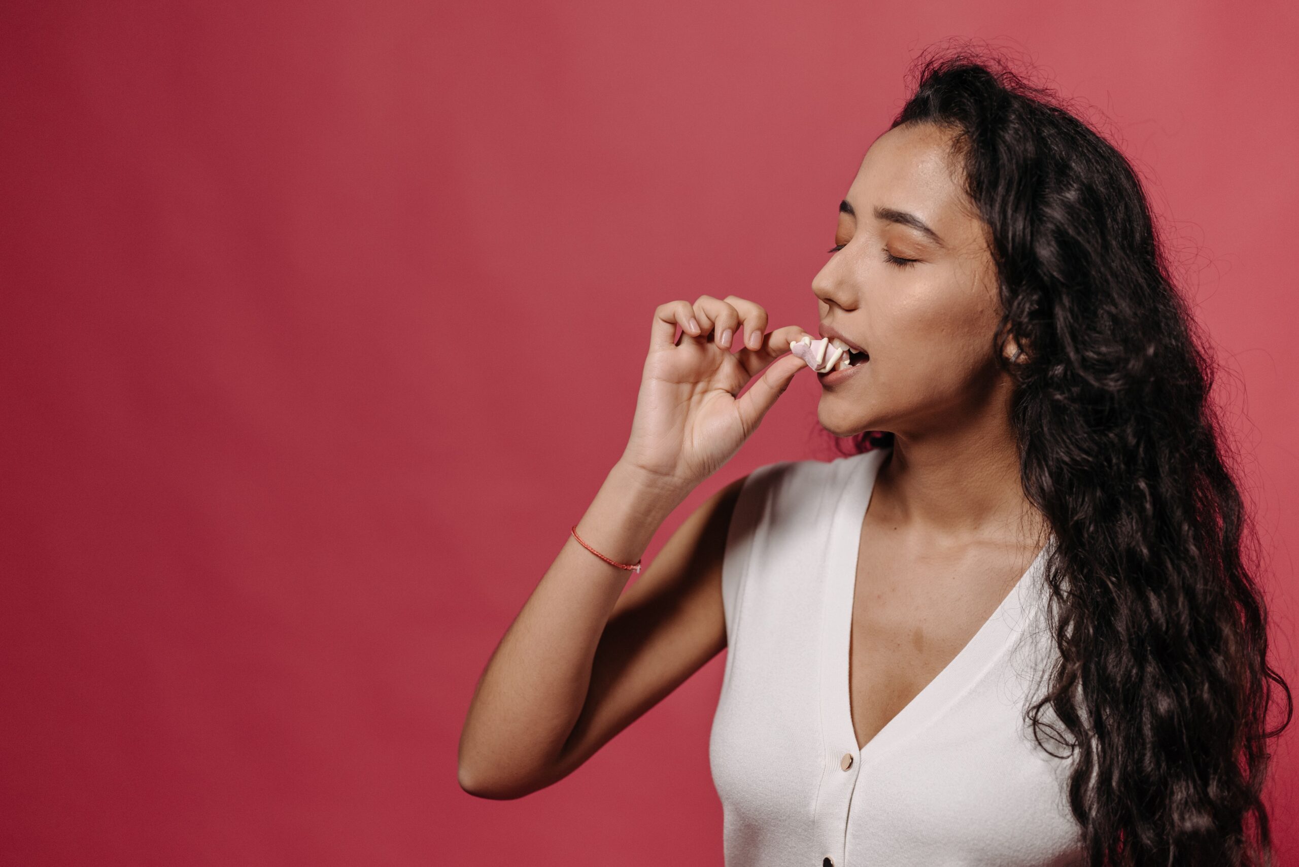 Stylish woman with curly hair savoring a treat against a pink studio backdrop.